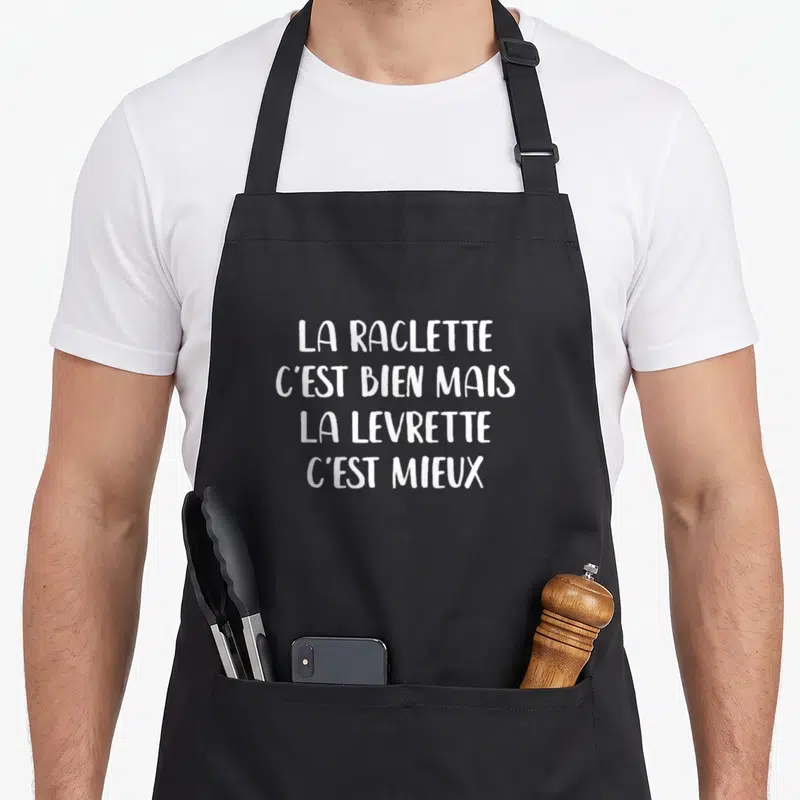 Tablier de cuisine noir avec le texte blanc La Raclette C'est Bien Mais La Levrette C'est Mieux imprimé au centre.