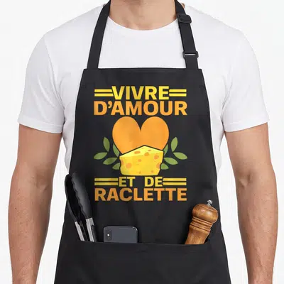 Tablier de cuisine noir avec un cœur orange, un bloc de fromage à trous et le texte vivre d'amour et de raclette en lettres jaunes.