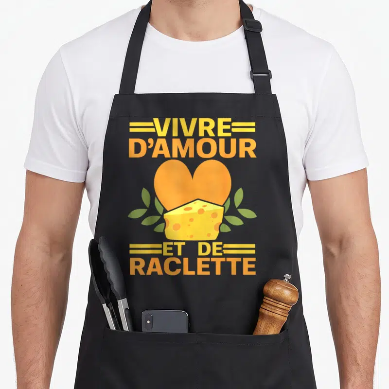 Tablier de cuisine noir avec un cœur orange, un bloc de fromage à trous et le texte vivre d'amour et de raclette en lettres jaunes.