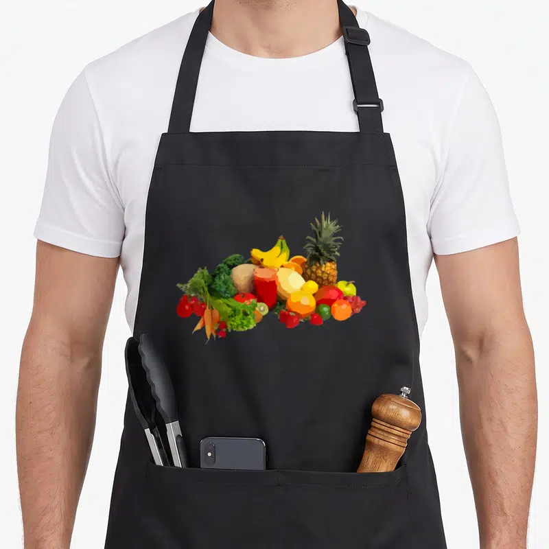 Tablier de cuisine noir avec une illustration colorée représentant un assortiment de fruits et légumes frais comme des bananes, un pineapple et des carottes.