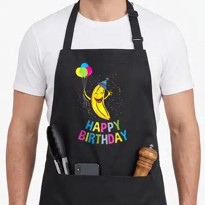 Tablier de cuisine noir avec une banane jaune souriante portant un chapeau de fête et tenant des ballons multicolores.
