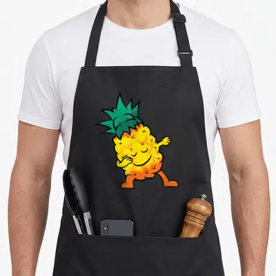 Tablier de cuisine noir avec illustration d'un ananas jaune aux pattes orange et couronne verte, semblant danser ou se tapoter le ventre.