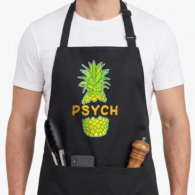 Tablier de cuisine noir avec illustration d'ananas jaune et vert coupé par le mot PSYCH en lettres capitales orange.