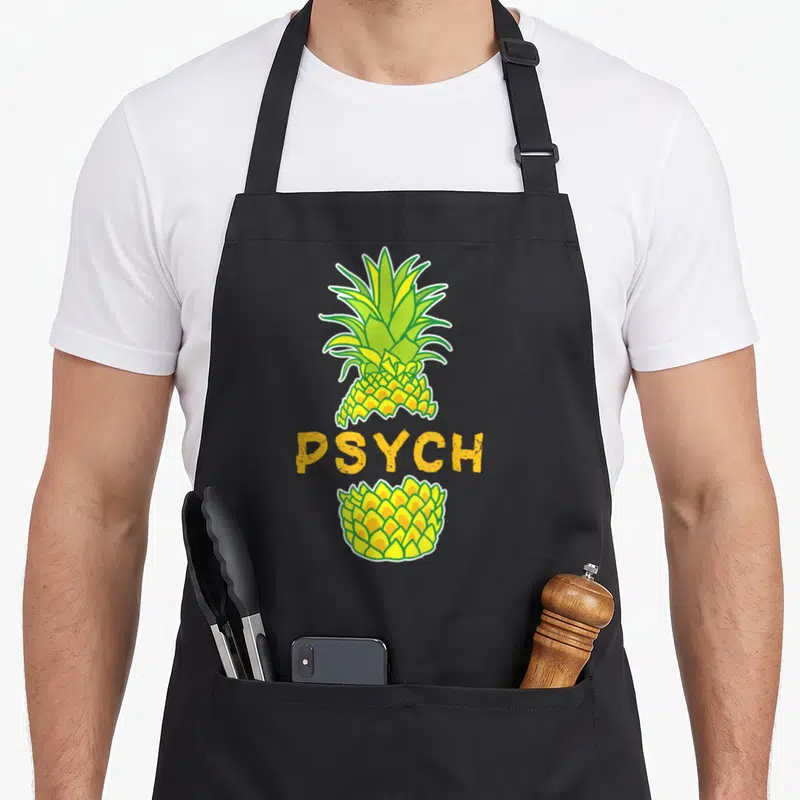 Tablier de cuisine noir avec illustration d'ananas jaune et vert coupé par le mot PSYCH en lettres capitales orange.