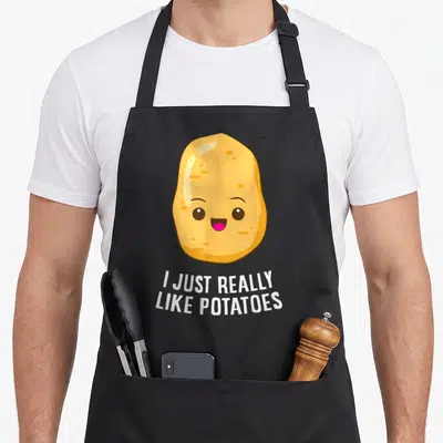 Tablier de cuisine noir avec une pomme de terre jaune souriante et un texte humoristique en anglais blanc.