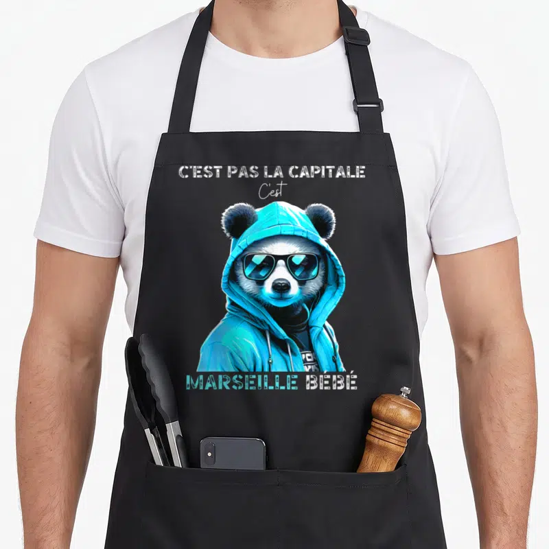 Tablier de cuisine noir avec illustration d'un panda en survêtement bleu et lunettes noires, arborant le texte C'est Pas la Capitale C'est Marseille bébé.