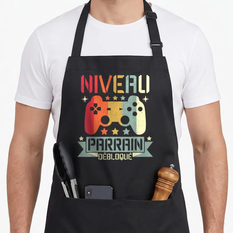 Tablier de cuisine noir avec illustration manette de jeu vidéo multicolore et texte Niveau Parrain Débloqué en typographie rétro.