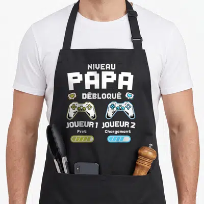 Tablier de cuisine noir avec inscription Niveau Papa Débloqué et deux manettes de jeu vidéo colorées.