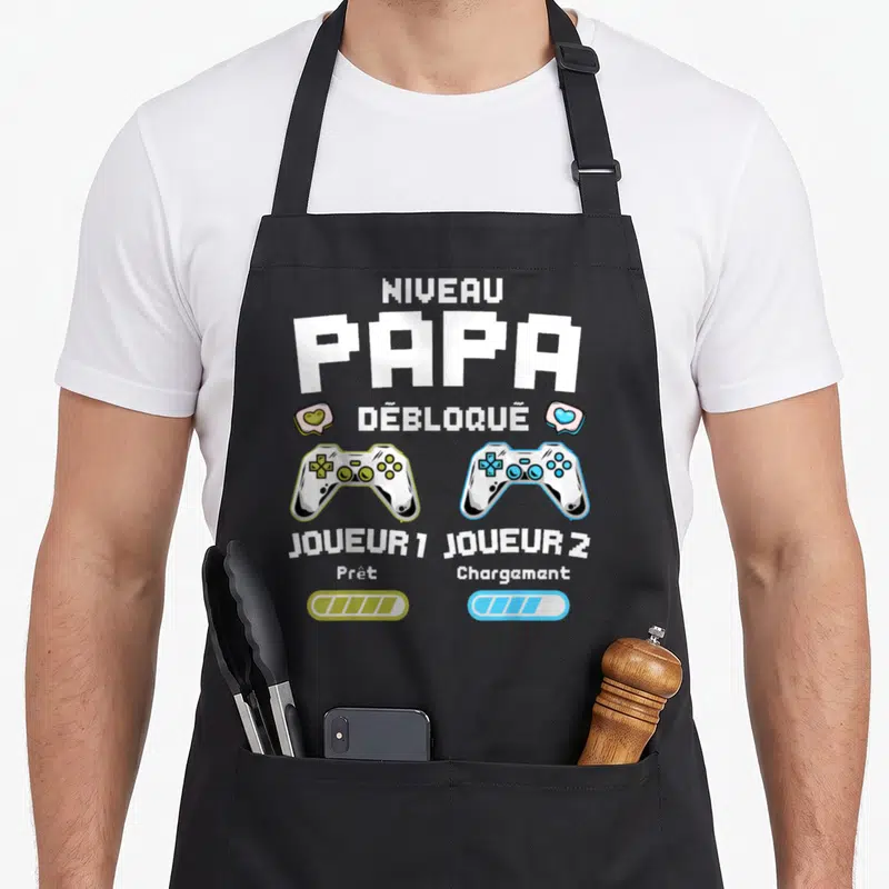 Tablier de cuisine noir avec inscription Niveau Papa Débloqué et deux manettes de jeu vidéo colorées.