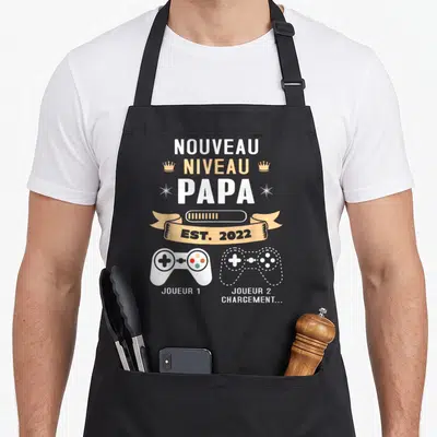 Tablier de cuisine noir avec inscription Nouveau Niveau Papa et deux manettes de jeu vidéo Joueur 1 et Joueur 2 chargement.