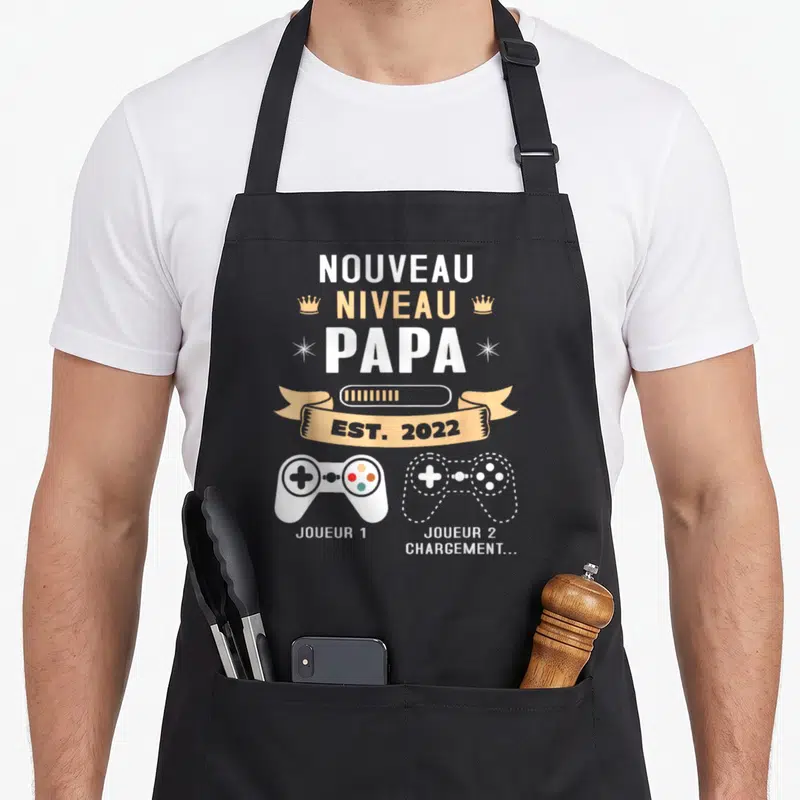 Tablier de cuisine noir avec inscription Nouveau Niveau Papa et deux manettes de jeu vidéo Joueur 1 et Joueur 2 chargement.