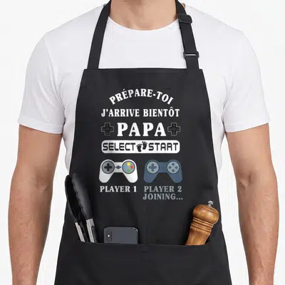 Tablier de cuisine noir avec texte blanc Prépare-toi Papa Select Start et deux manettes de jeu vidéo colorées.