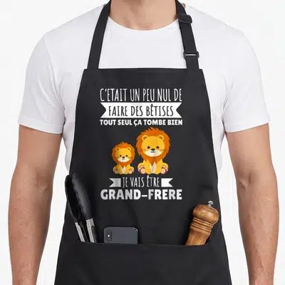 Tablier de cuisine noir avec deux lions et le texte C'était un peu nul de faire des bêtises tout seul ça tombe bien je vais être grand-frère
