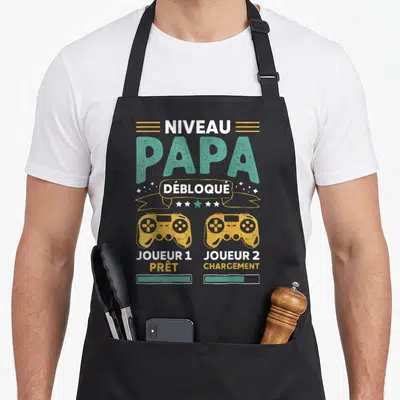 Tablier de cuisine noir avec illustration niveau papa débloqué deux manettes jaunes et texte joueur 1 prêt joueur 2 chargement en typographie gaming.