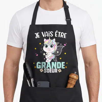 Tablier de cuisine noir avec une licorne rose et l'annonce Je vais être grande soeur entourée d'étoiles.