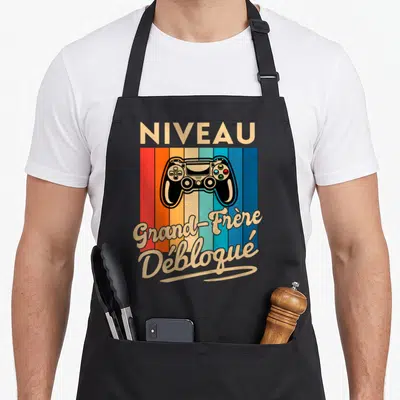 Tablier de cuisine noir avec illustration de manette de jeu vidéo et texte Niveau Grand Frère Débloqué sur fond coloré vintage.
