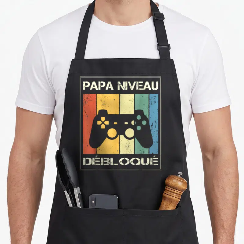 Tablier de cuisine noir avec illustration vintage manette de jeu vidéo et texte Papa niveau débloqué.