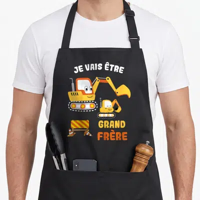 Tablier de cuisine noir avec deux pelles mécaniques jaunes souriantes et le texte Je vais être grand frère 2026.