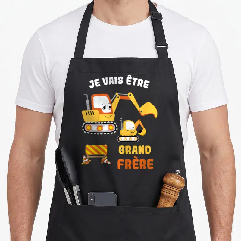 Tablier de cuisine noir avec deux pelles mécaniques jaunes souriantes et le texte Je vais être grand frère 2026.