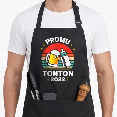 Tablier de cuisine noir avec motif Promu Tonton 2022 illustrant un biberon et une bière sur fond de soleil rétro.