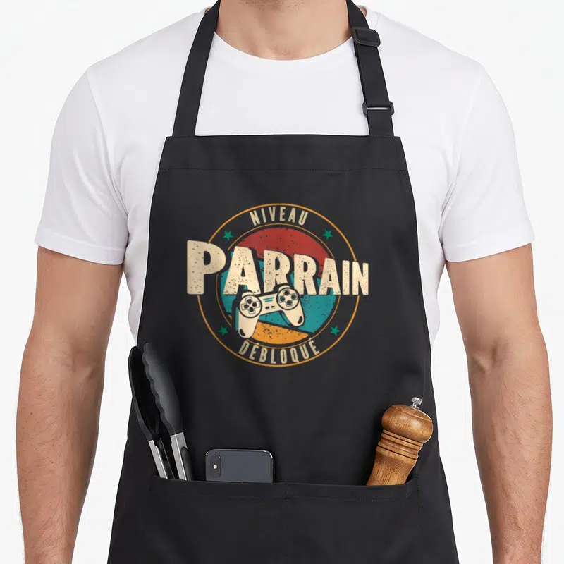 Tablier de cuisine noir avec logo Niveau Parrain Débloqué et manette de jeu vidéo colorée.