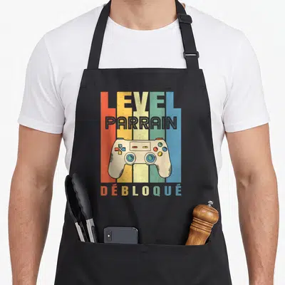 Tablier de cuisine noir avec illustration manette de jeu vidéo et texte Level Parrain Débloqué sur bandes colorées verticales.