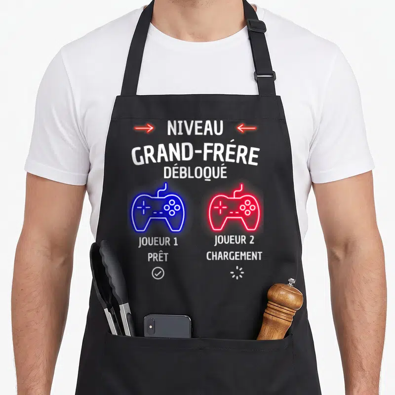 Tablier de cuisine noir avec texte Niveau Grand-Frère Débloqué et deux manettes de jeu vidéo bleue et rouge.
