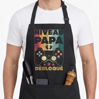 Tablier de cuisine noir avec inscription Niveau Papa Débloqué, manette de jeu vidéo et traces de pas colorées sur fond déstructuré.