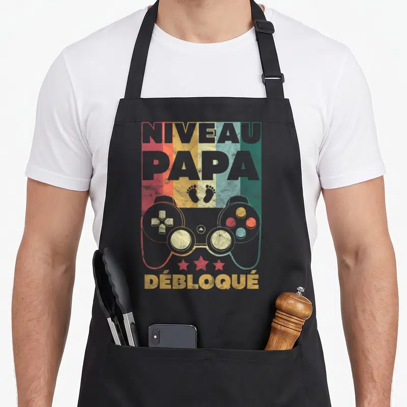 Tablier de cuisine noir avec inscription Niveau Papa Débloqué, manette de jeu vidéo et traces de pas colorées sur fond déstructuré.