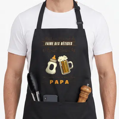 Tablier de cuisine noir avec illustration d'un biberon et d'une bière et texte humoristique sur la paternité.