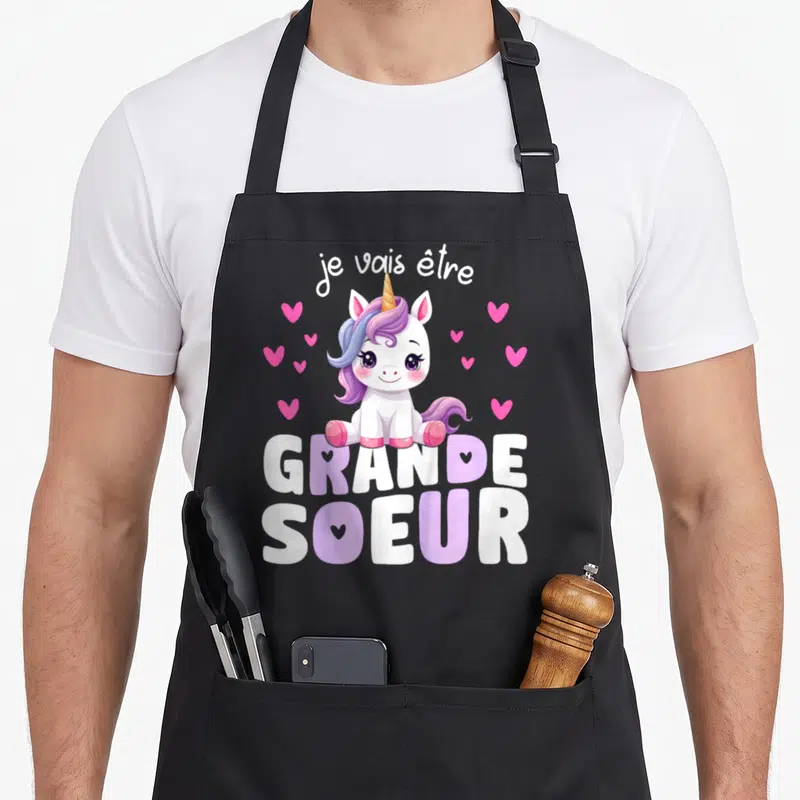 Tablier de cuisine noir avec licorne rose et texte Je vais être Grande Soeur entouré de cœurs roses.