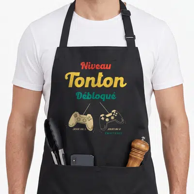 Tablier de cuisine noir avec inscription Niveau Tonton Débloqué et deux manettes de jeu vidéo Joueur 1 et Joueur 2 en attente.