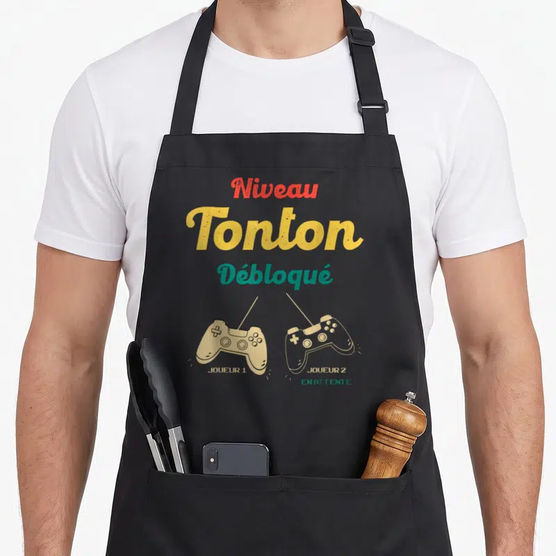 Tablier de cuisine noir avec inscription Niveau Tonton Débloqué et deux manettes de jeu vidéo Joueur 1 et Joueur 2 en attente.