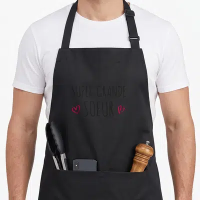 Tablier de cuisine noir avec l'inscription Super Grande Soeur et deux cœurs roses stylisés sur le devant.