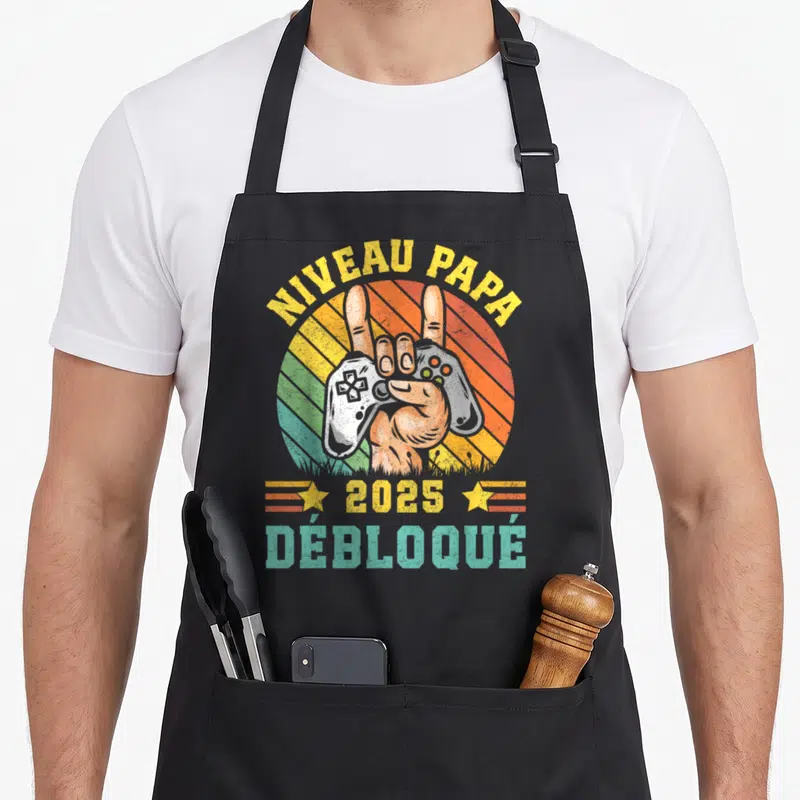 Tablier de cuisine noir avec une main tenant une manette et le texte Niveau Papa Débloqué 2025 en style rétro.