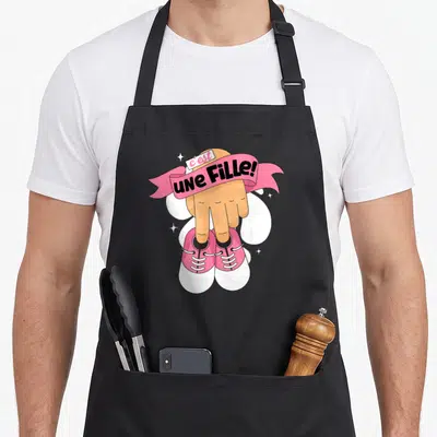 Tablier de cuisine noir avec une main pointant vers des chaussures roses et le texte C'est une Fille.