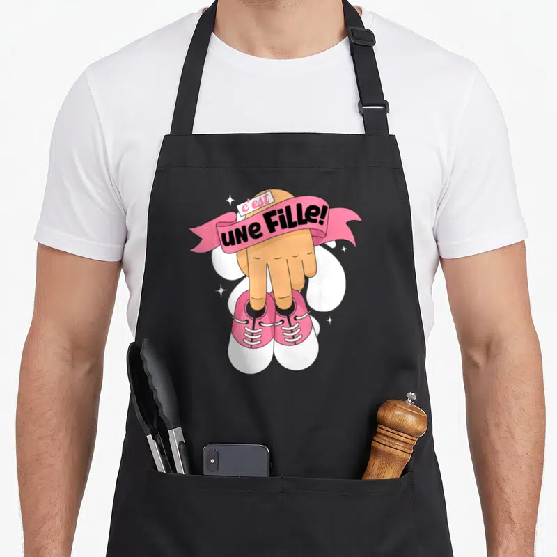 Tablier de cuisine noir avec une main pointant vers des chaussures roses et le texte C'est une Fille.
