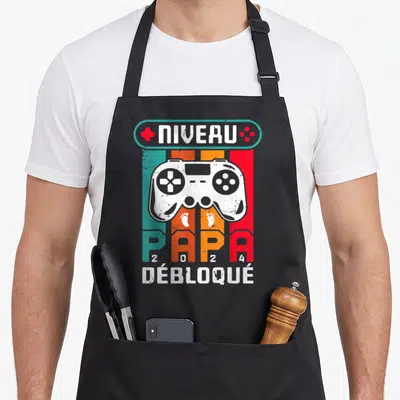 Tablier de cuisine noir avec un manette de jeu vidéo et l'inscription Niveau Papa 2024 Débloqué sur fond rayé coloré.