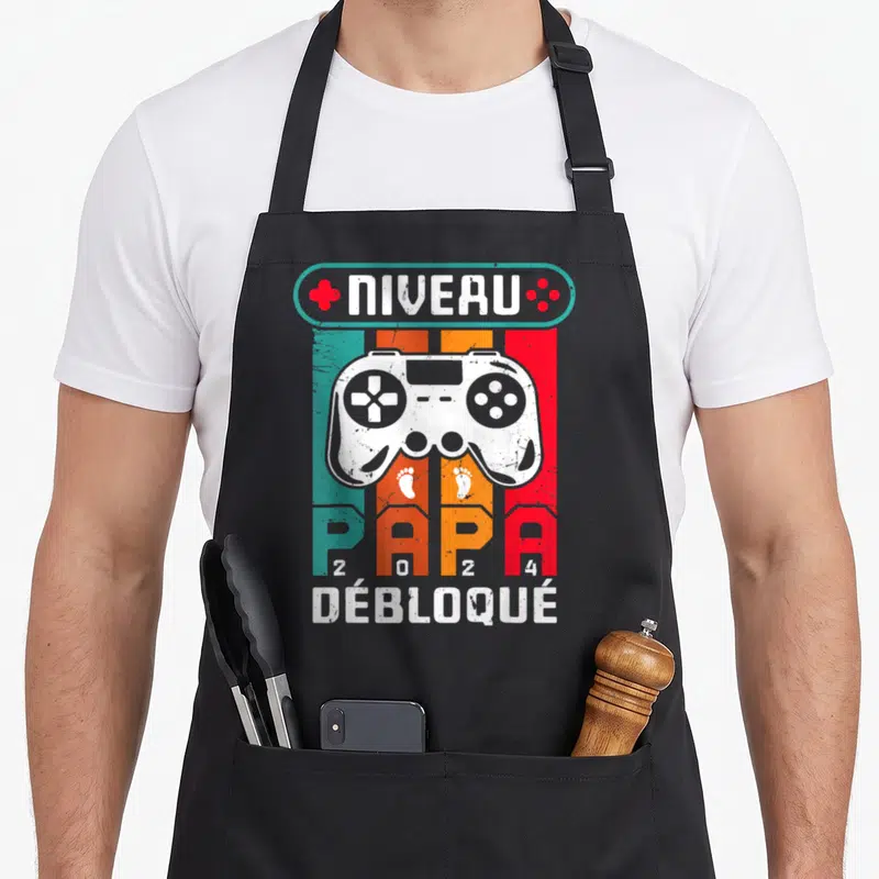Tablier de cuisine noir avec un manette de jeu vidéo et l'inscription Niveau Papa 2024 Débloqué sur fond rayé coloré.