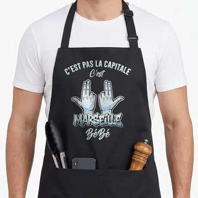 Tablier de cuisine noir avec deux mains marseillaises stylisées et le texte C'est pas la capitale Marseille Bébé en typographie blanche et bleue.