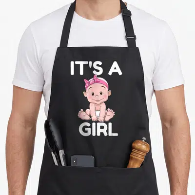 Tablier de cuisine noir avec illustration « It's a Girl » et bébé souriant coiffé d'un ruban rose à pois blancs.