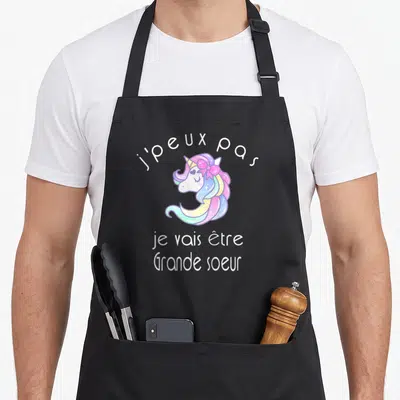 Tablier de cuisine noir avec une licorne colorée et le texte j'peux pas je vais être Grande soeur imprimé en blanc.