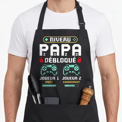 Tablier de cuisine noir avec inscription Niveau Papa 2023 Débloqué et deux manettes de jeu vidéo vertes.