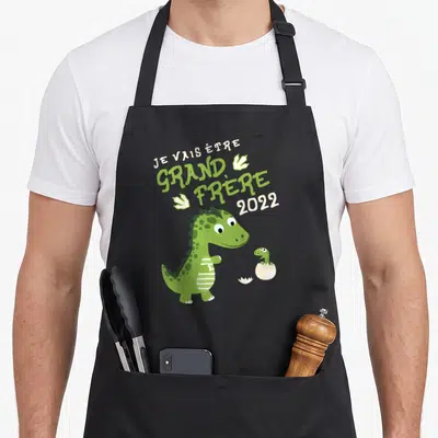 Tablier de cuisine noir avec un dinosaure vert et le texte Je vais être grand frère 2022 imprimé au centre.