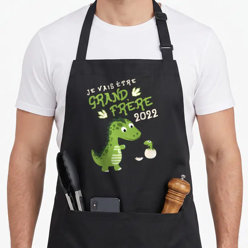 Tablier de cuisine noir avec un dinosaure vert et le texte Je vais être grand frère 2022 imprimé au centre.