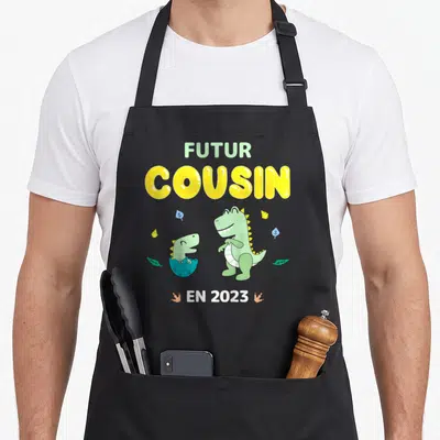 Tablier de cuisine noir avec inscription Futur Cousin En 2023 et deux dinosaures verts mignons sur fond uni.