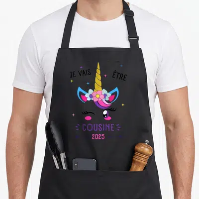 Tablier de cuisine noir avec illustration de licorne colorée et texte announcing une future cousine pour l'année 2025.