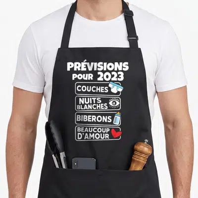 Tablier de cuisine noir avec l'annonce Prévisions pour 2023 Couches Nuits blanches Biberons Beaucoup d'amour illustrée.