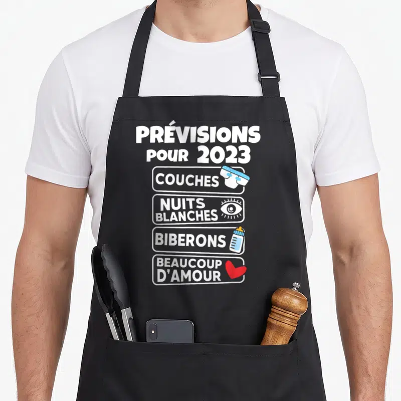 Tablier de cuisine noir avec l'annonce Prévisions pour 2023 Couches Nuits blanches Biberons Beaucoup d'amour illustrée.
