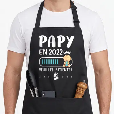 Tablier de cuisine noir avec le texte Papy en 2022 Veuillez patienter et un bébé tenant une bouteille.